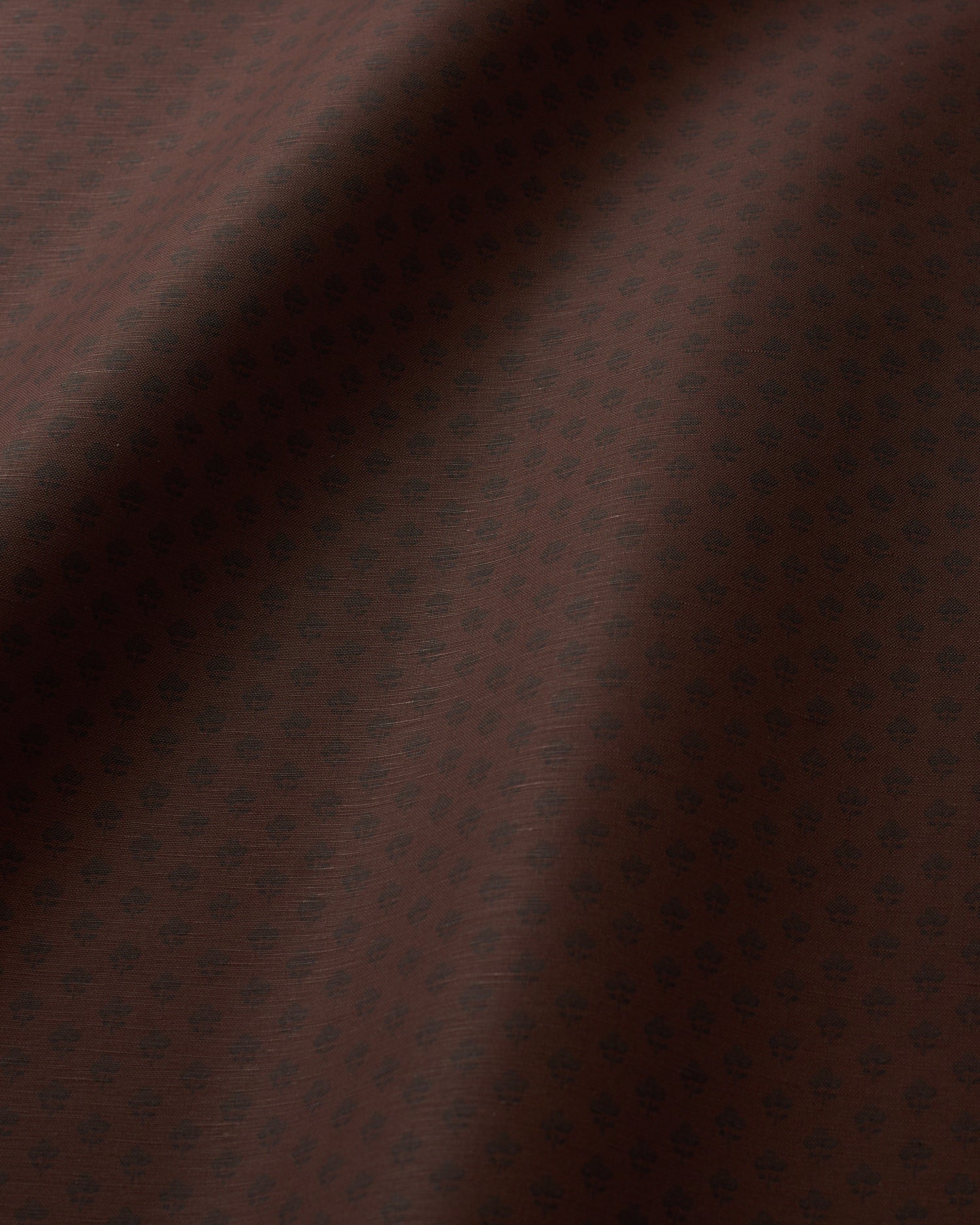 A close-up of dark brown fabric with a subtle repeating small floral or medallion pattern and soft folds creating shadows.