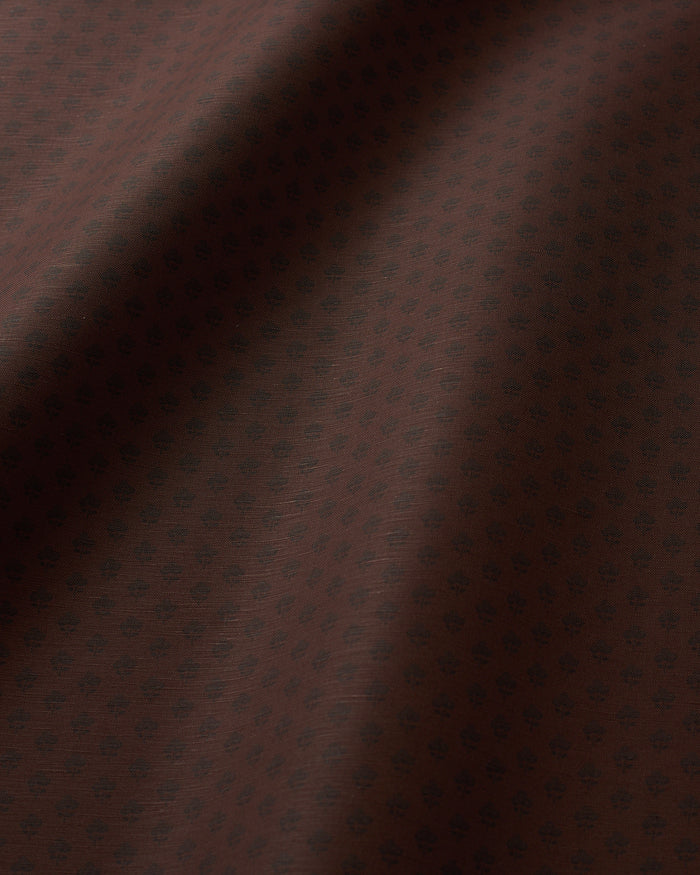 A close-up of dark brown fabric with a subtle repeating small floral or medallion pattern and soft folds creating shadows.