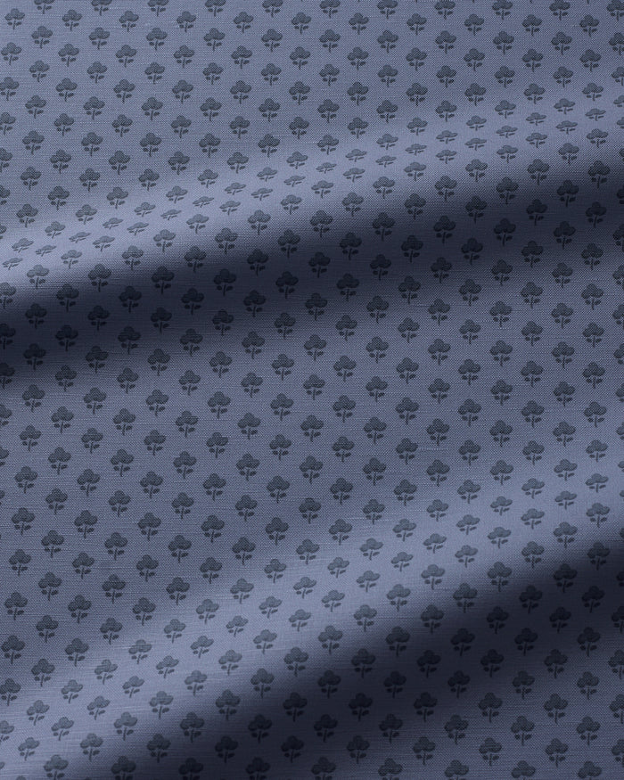 A close-up of a fabric surface with a repeating small floral pattern in a muted blue-gray tone, with soft diagonal shadows across the material.